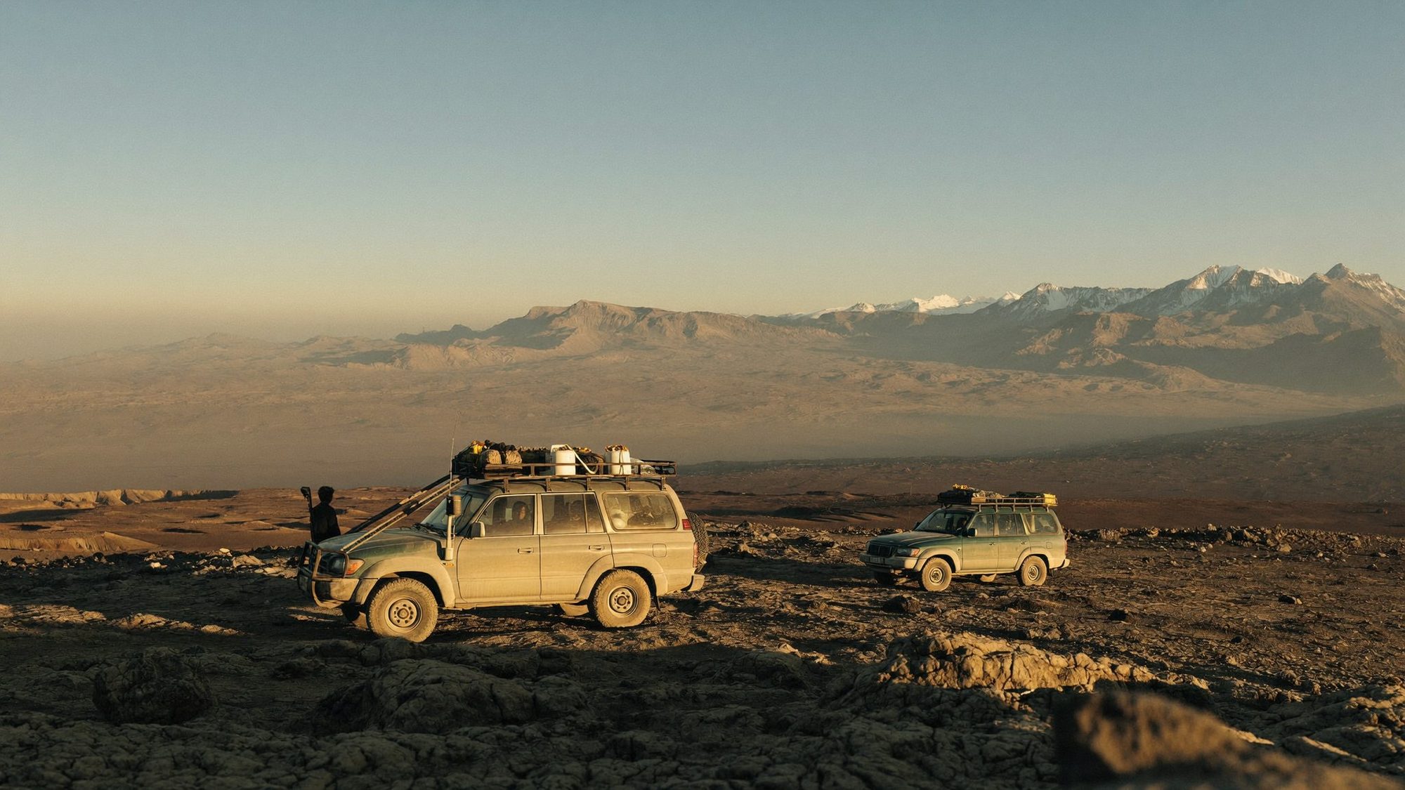 Two 4x4 vehicles on a high plateau below snowy mountains