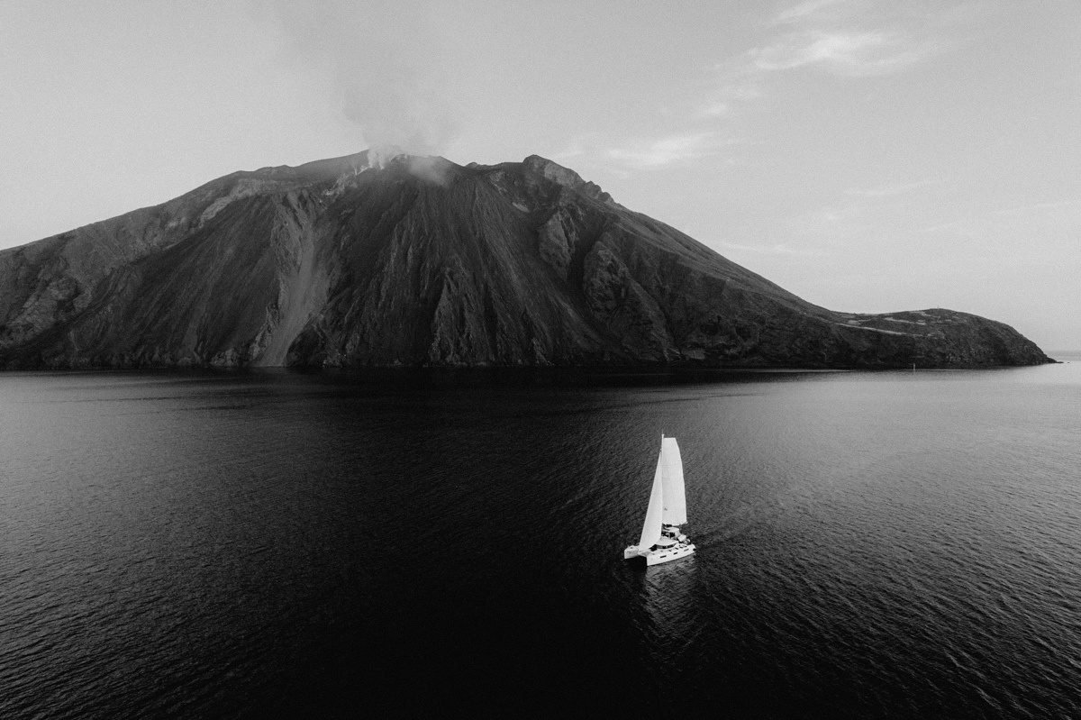 A catamaran sailing below the Stromboli volcano, black and white